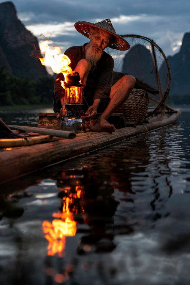 A cormorant fisherman lighting his lantern on a bamboo raft in Guilin at sunset.