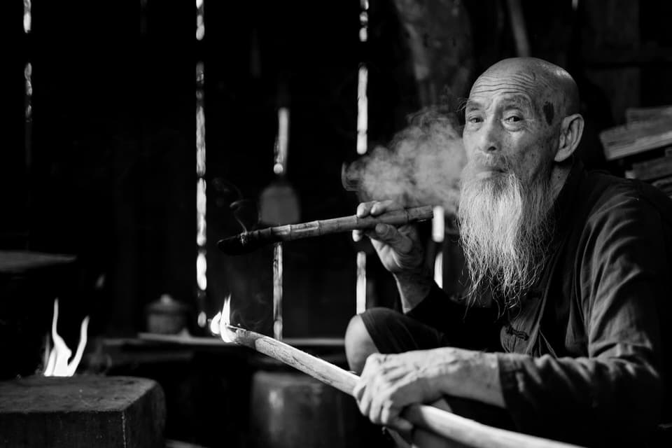 Black-and-white portrait of a cormorant fisherman smoking a pipe at home in Guilin, China.